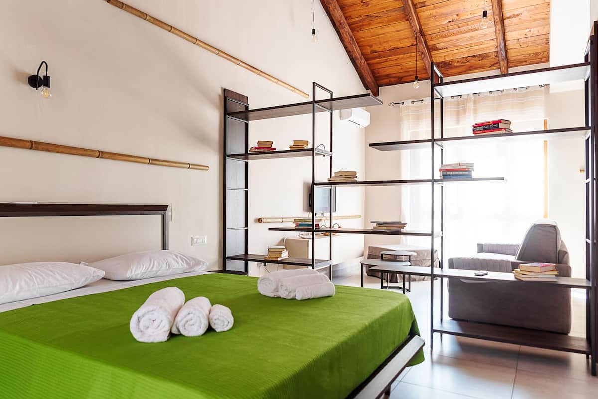 A modern hotel room with a green bed, white pillows, a wooden ceiling, and built-in shelves with books.