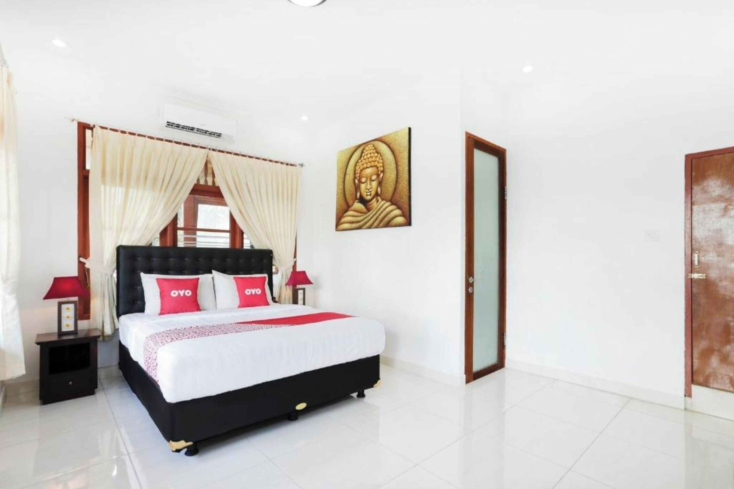 A bedroom with a black headboard, white bedding, and red pillows. There is a wall-mounted air conditioning unit, a framed picture, and a door with a glass panel.