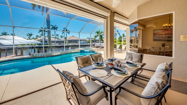 Un espace repas aménagé au bord de la piscine, offrant une vue sur la piscine et les palmiers, avec un ventilateur de plafond et une table à manger dressée avec des assiettes et des verres.