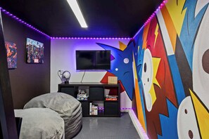 Una habitación con un gran mural en la pared, un televisor de pantalla plana, un sillón informal y una estantería con diversos objetos.