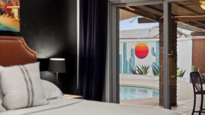 Une chambre d’hôtel comprenant un lit, une table de chevet, une lampe et une vue sur un espace extérieur avec piscine, agrémenté d’une fresque murale.