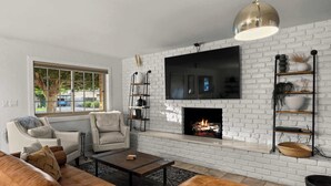 Un salon moderne avec une cheminée en briques blanches, un téléviseur à écran plat fixé au-dessus, et un coin salon confortable avec un canapé en cuir marron et un fauteuil blanc.