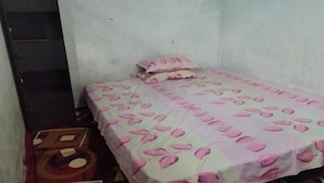 Una habitación pequeña con una cama individual, una colcha estampada y una almohada. Se ve un armario a la izquierda y una pequeña mesa con un objeto decorativo en el suelo.