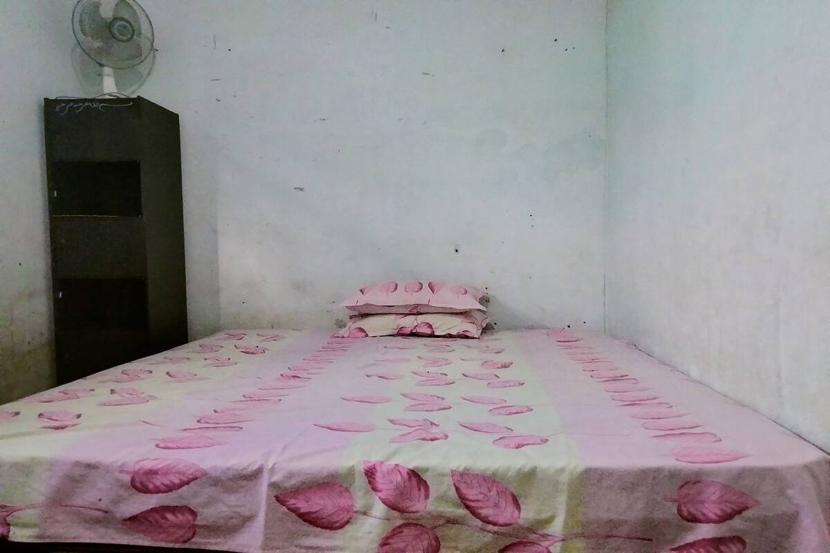 Une petite pièce simple, avec un lit recouvert d’une couverture à motifs floraux roses, un seul oreiller et un ventilateur fixé au mur.