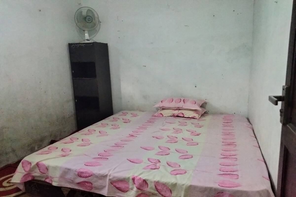 Une petite chambre avec un lit simple, une couvre-lit à motifs floraux roses, une table de chevet et un ventilateur sur pied.