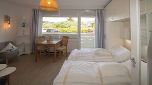 A bedroom with two beds, a dining table with chairs, a small sofa, and a window with a view of the outside. - Die Wohnung Treibholz mit Balkon Liegt in Einer Sehr Ruhigen Lage in Hörnum (Hörnum (Sylt))