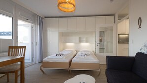 A compact room with two single beds, a dining table, a sofa, and a kitchen area visible through an open door. - Die Wohnung Treibholz mit Balkon Liegt in Einer Sehr Ruhigen Lage in Hörnum (Hörnum (Sylt))