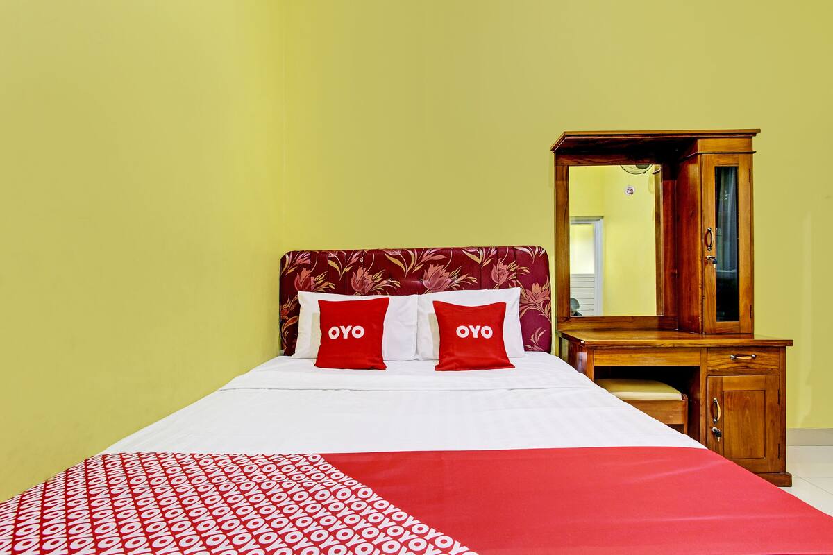 Un lit recouvert d’une couvre-lit à motifs rouges et blancs, deux oreillers ornés du mot « OYO », une table de chevet en bois et un miroir.