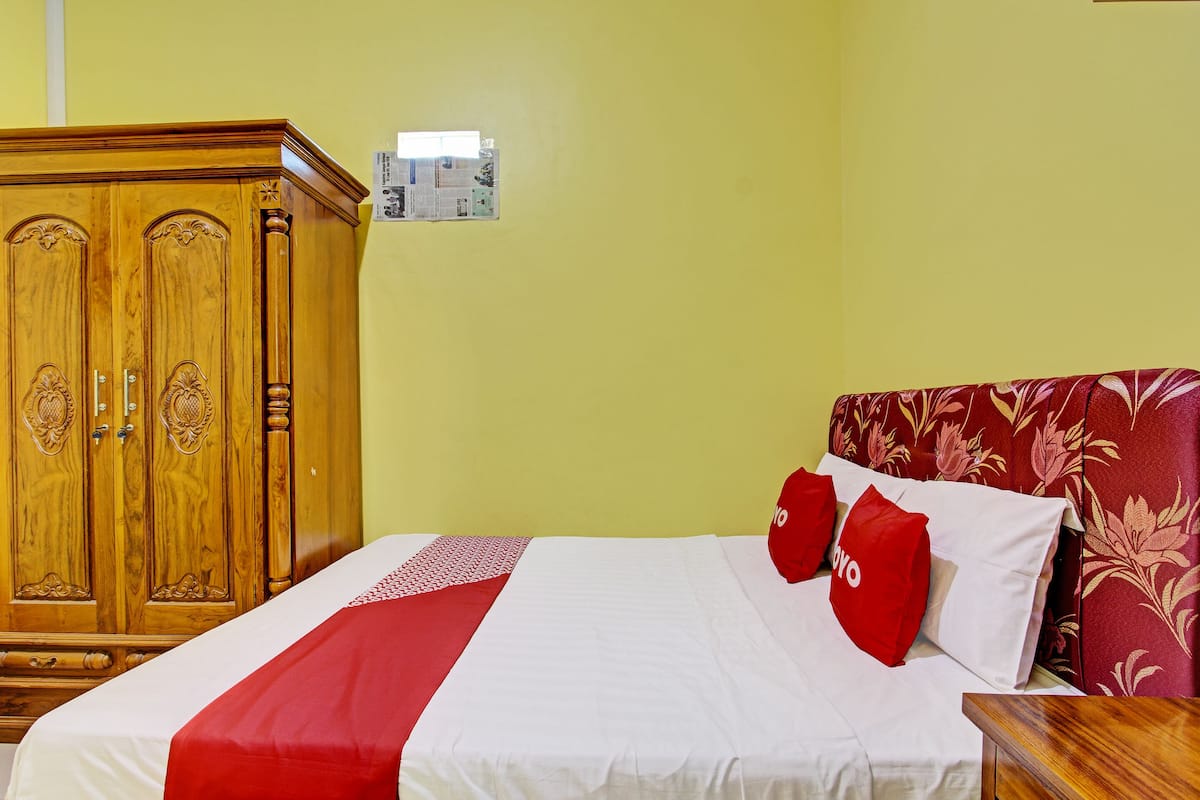Une chambre d’hôtel avec une armoire en bois, un lit avec une literie rouge et blanche, et une tête de lit à motifs floraux.