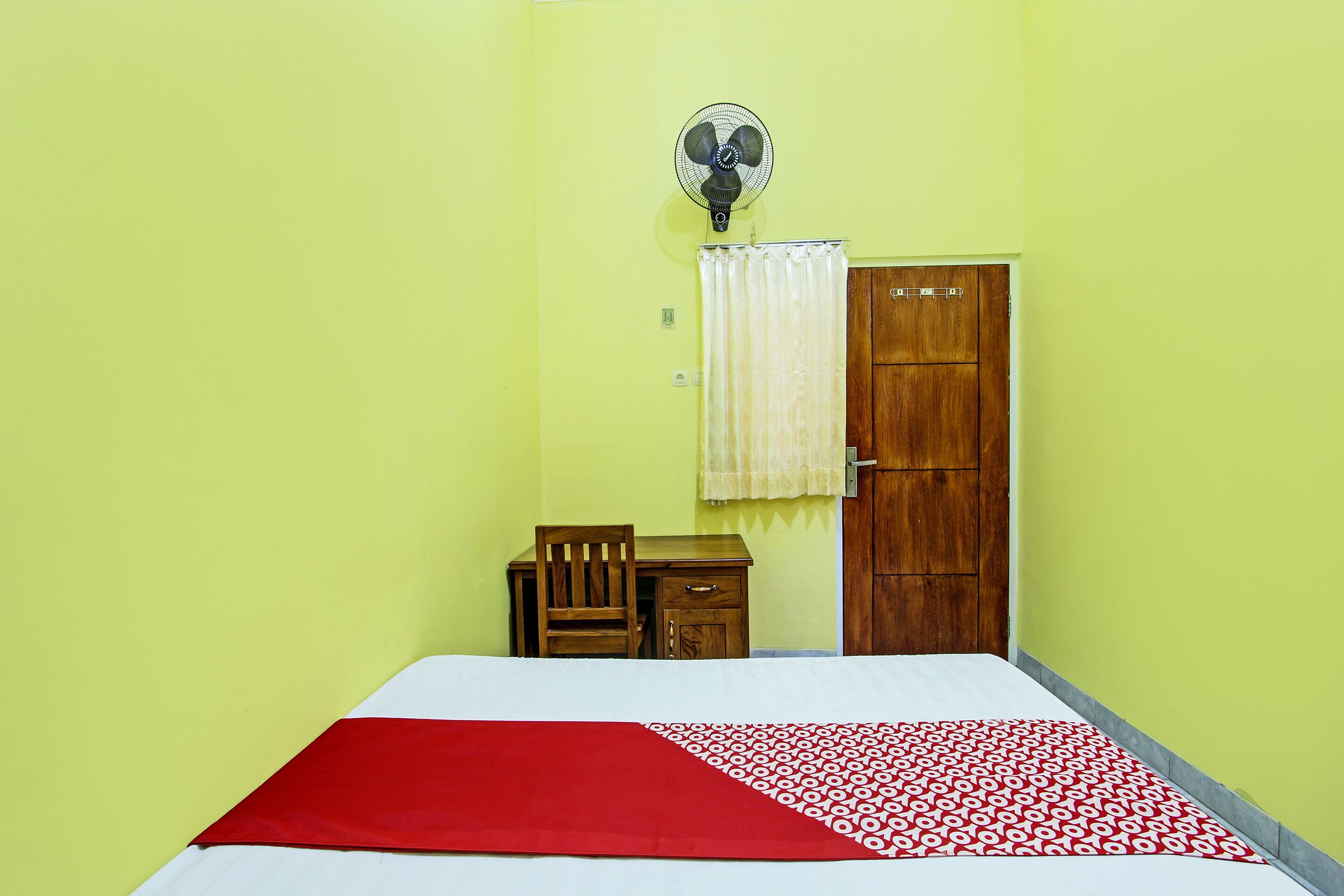 A room with a bed, a wooden desk, a chair, a wooden wardrobe, and a wall-mounted fan.