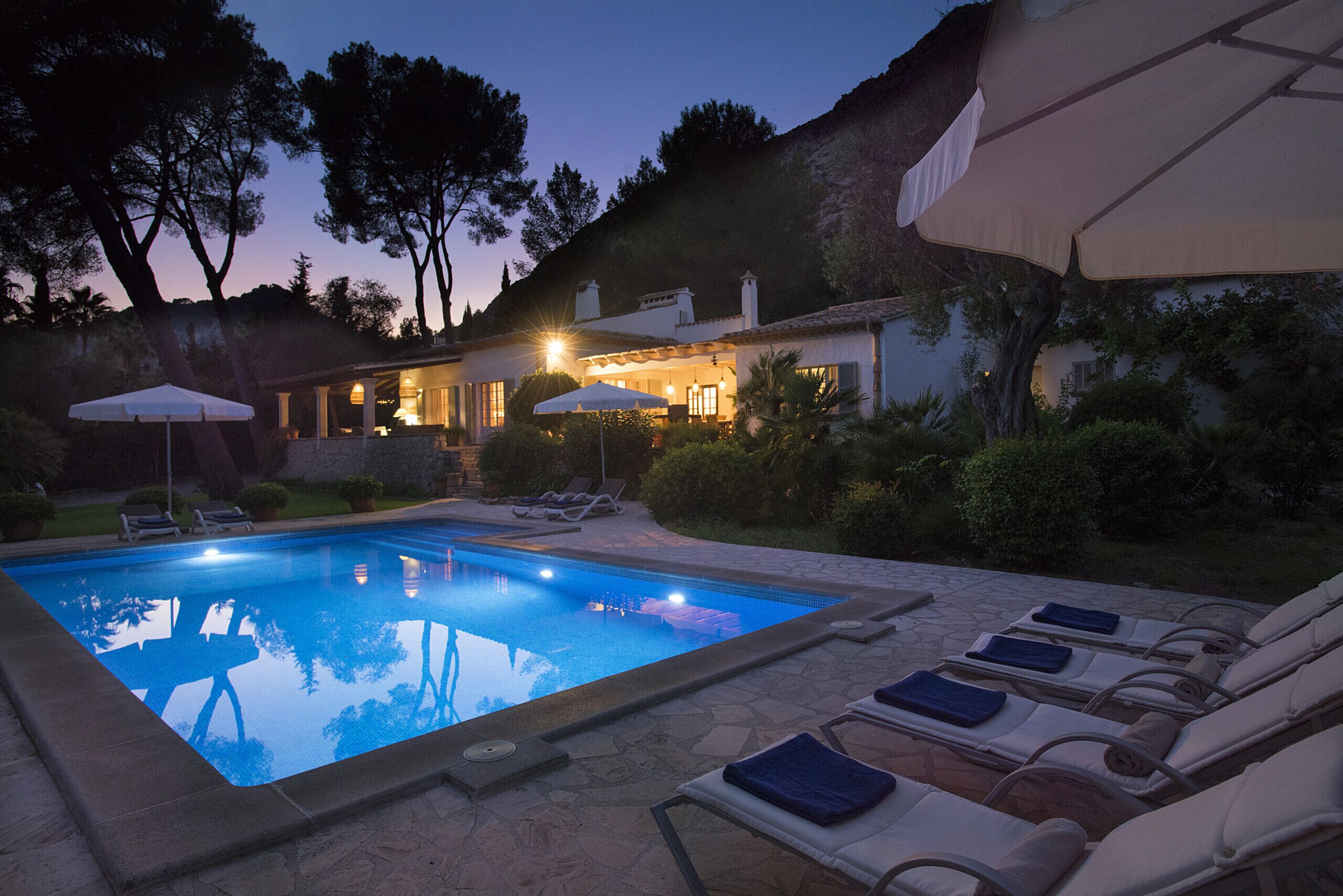 Uma piscina com espreguiçadeiras e guarda-sóis, cercada por árvores e um edifício iluminado.