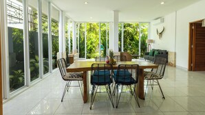 A spacious dining area with a large wooden table and metal chairs, surrounded by large windows offering a view of lush greenery. - Casa Sweet Lips by Betterplace (Canggu)