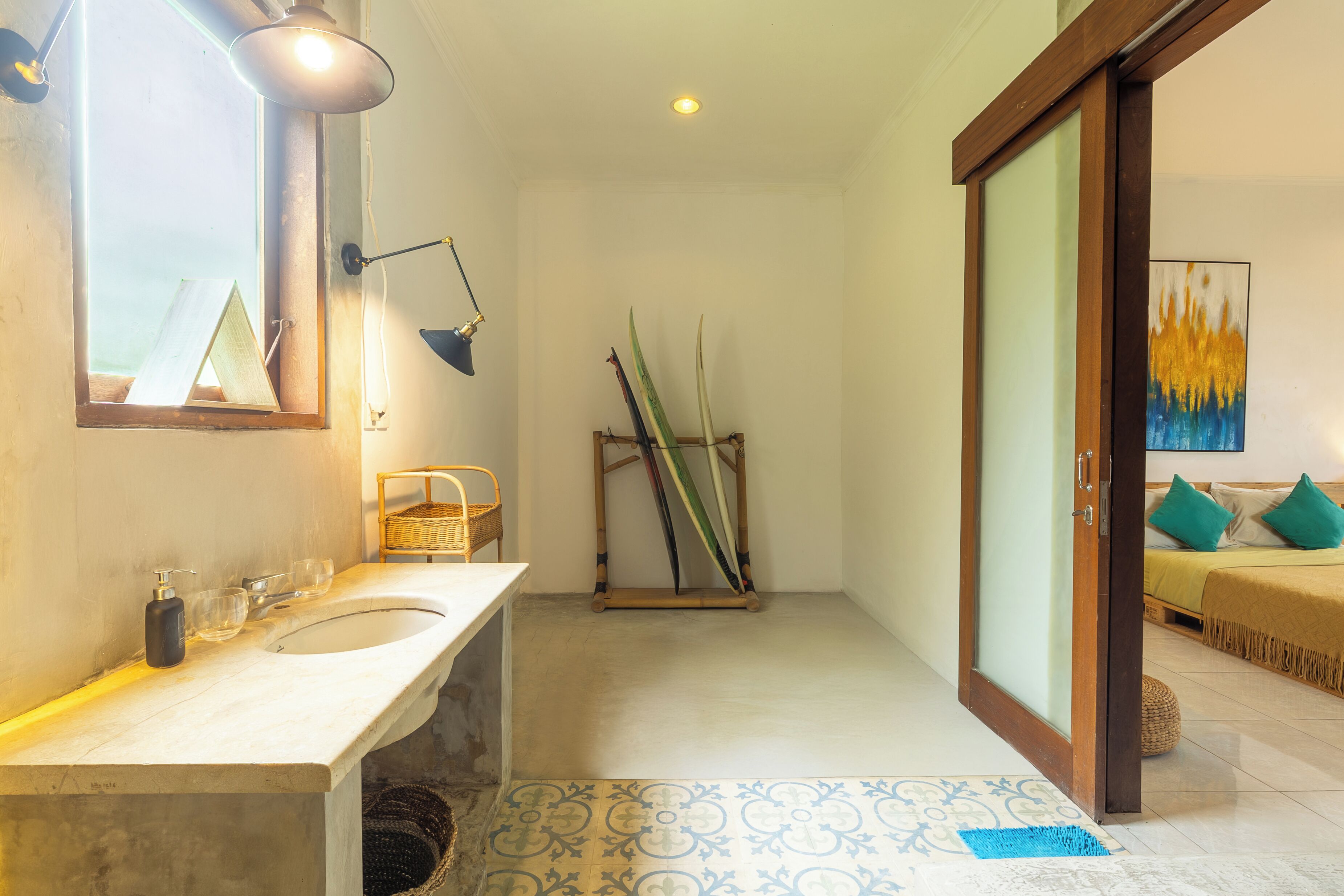 Un baño con un espejo grande, un lavabo y una ventana. En la habitación contigua, hay una cama con almohadas y un soporte de madera con utensilios de limpieza.