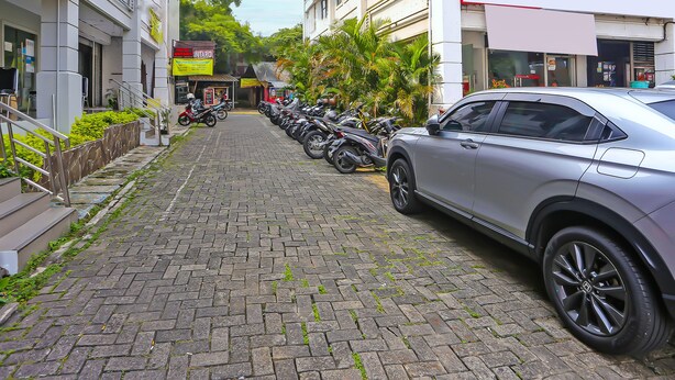 Une rue pavée bordée de motos et de voitures garées, des bâtiments avec des enseignes et un escalier menant à l’entrée d’un immeuble.