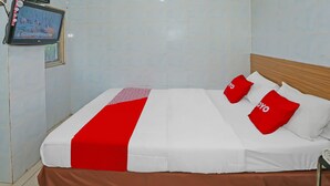 Un lit bien fait, avec une literie blanche et rouge, un couvre-lit marron et deux oreillers rouges ornés d’un motif blanc. Un téléviseur est fixé au mur au-dessus du lit.