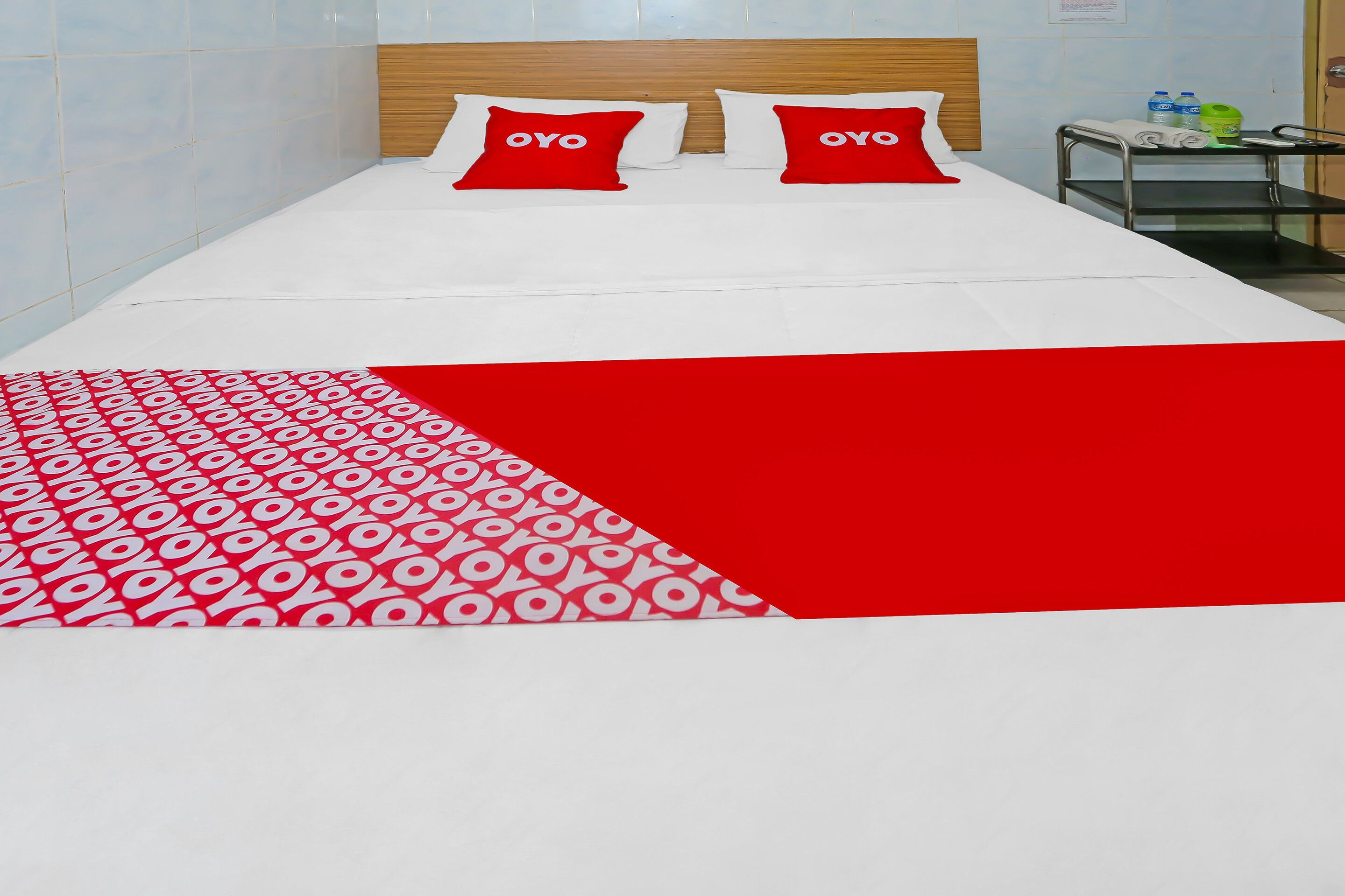 A hotel room with a bed, two red pillows with "OYO" text, a red and white patterned throw, and a small table with toiletries.
