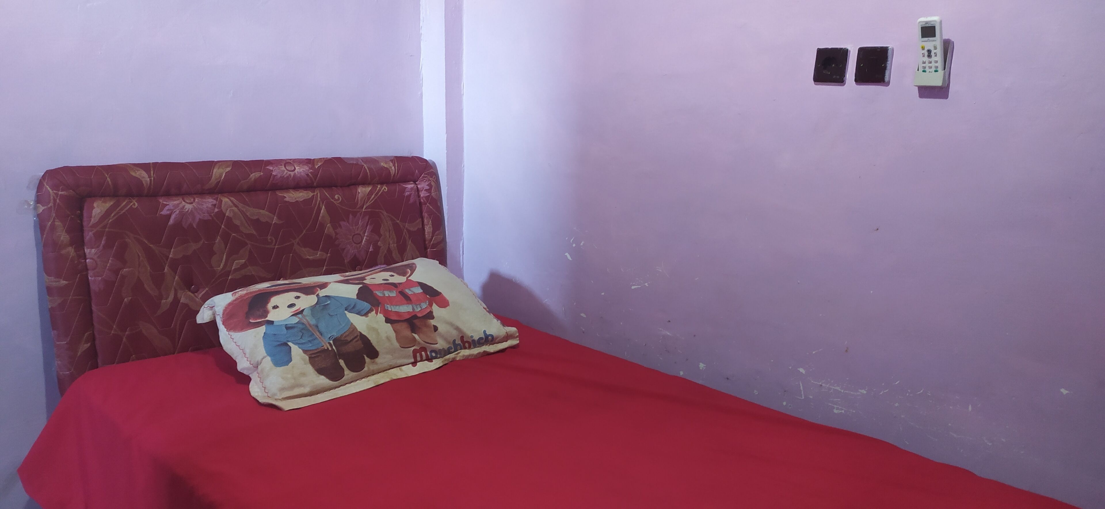 A small room with a single bed, a red bedspread, a pillow with cartoon characters, and a wall-mounted phone.