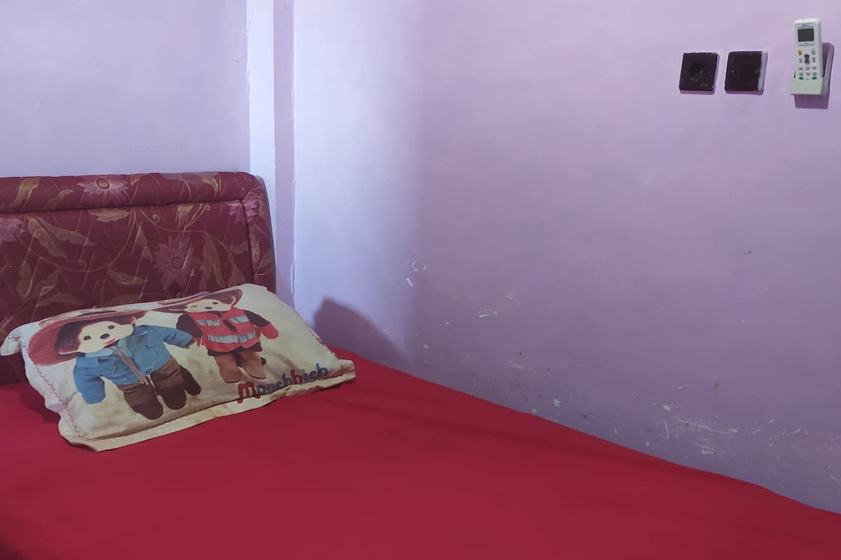 Une petite pièce avec un lit simple, une couverture rouge, un oreiller orné de personnages de dessins animés et un téléphone fixé au mur.