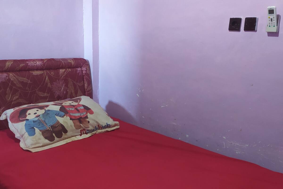 A small room with a single bed, a red bedspread, a pillow with cartoon characters, and a wall-mounted phone.