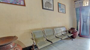 Een wachtruimte met metalen stoelen, een grote keramieken pot en ingelijste foto's aan de muur.