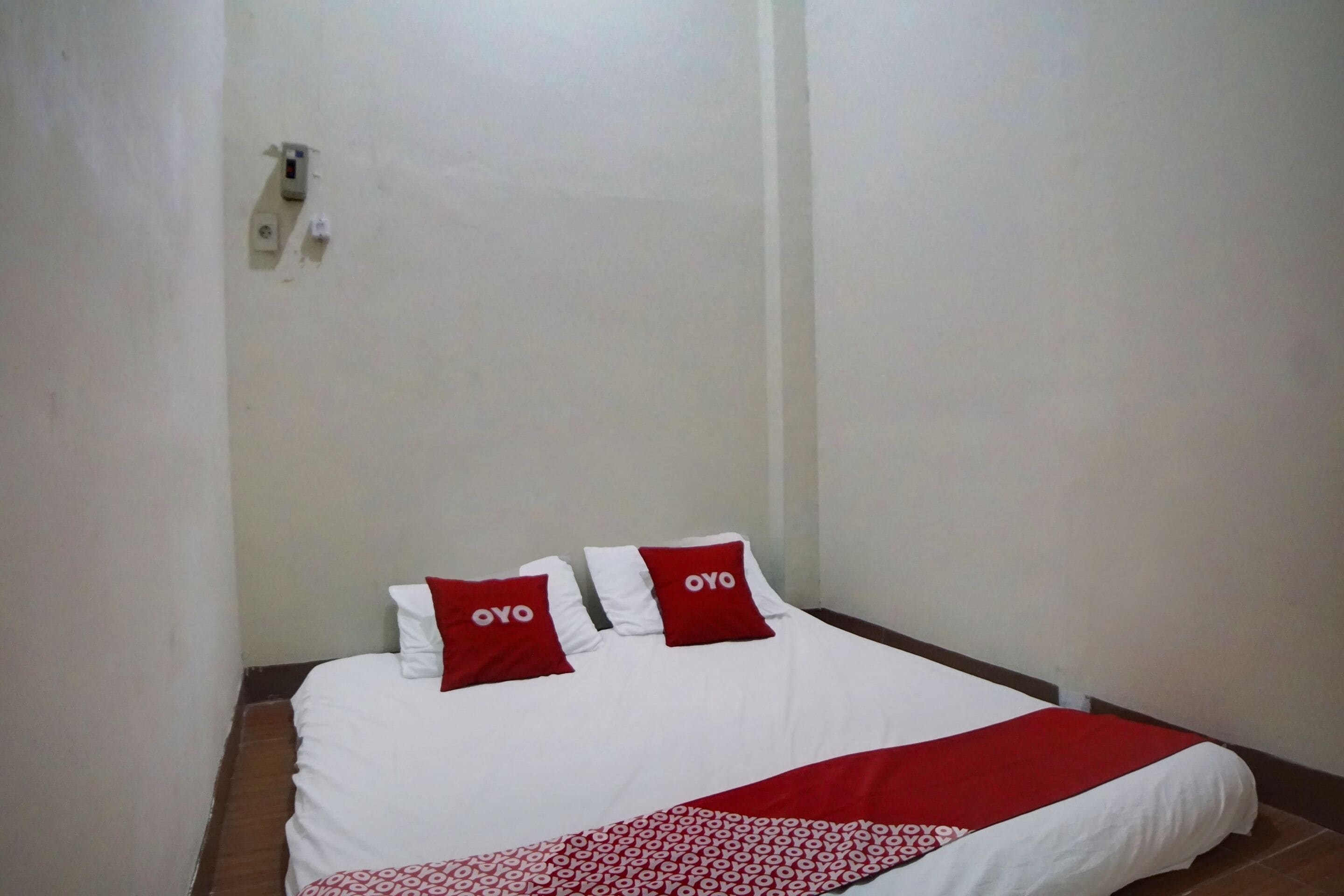 A neatly made bed with white bedding and red pillows, a red and white patterned throw blanket, and a wall-mounted electrical outlet.