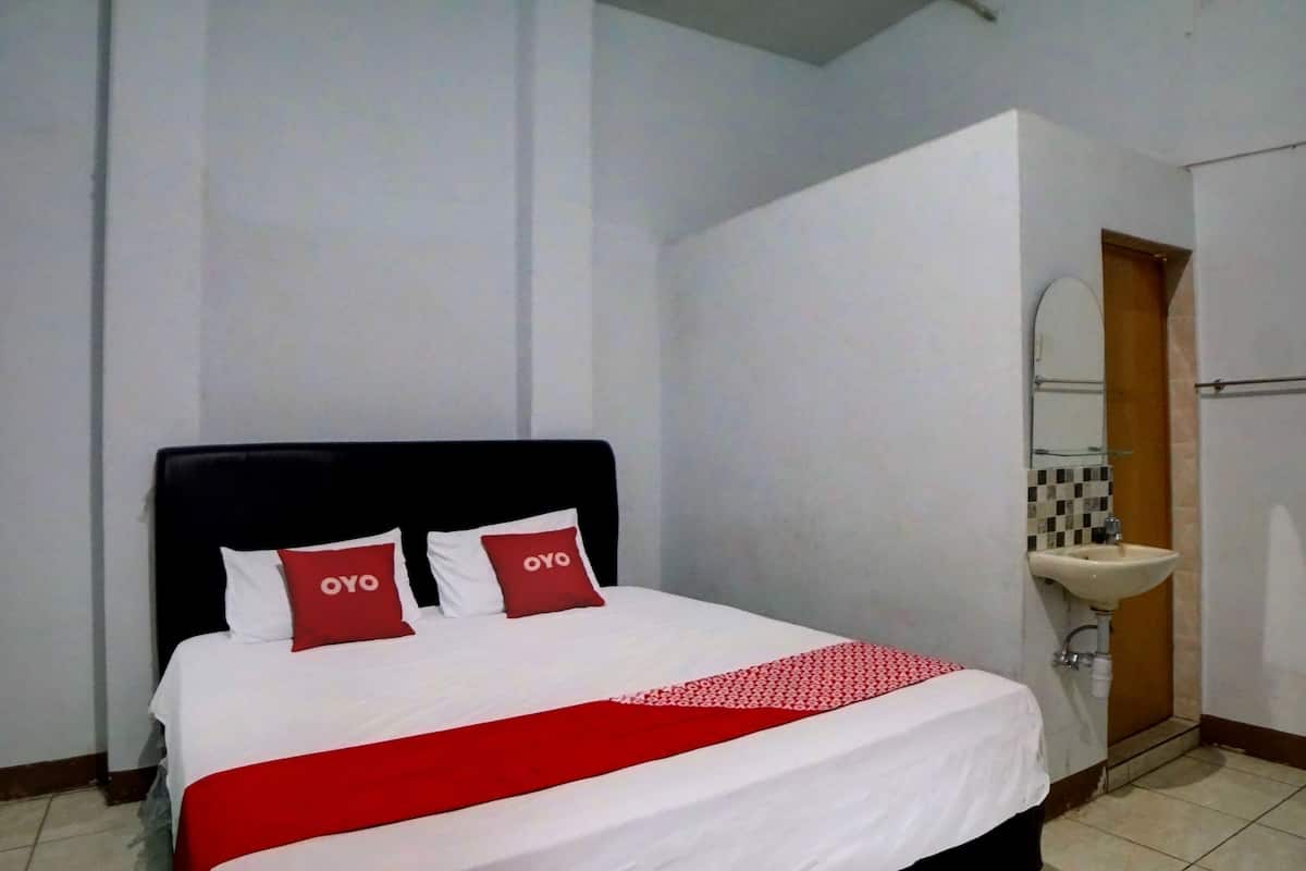 Ein Hotelzimmer mit Bett, zwei Kissen, einer rot-weißen Bettdecke, einer weißen Wand, einer Holztür und einem Badezimmer mit Waschbecken und Spiegel.