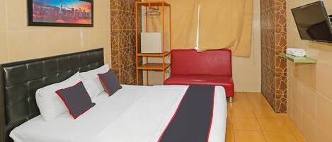 A hotel room with a bed, a TV, a red chair, a shelf with a remote, and a painting on the wall.