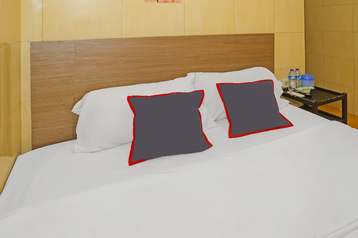 A neatly made bed with white sheets and two pillows, one white and one grey with red trim, against a wooden headboard. A small table beside the bed holds a remote control, a water bottle, and a blue cup.