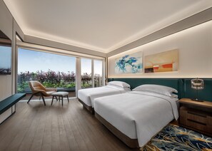 Et moderne hotelværelse med to senge, et siddeområde med en stol, et skrivebord og et stort vindue med udsigt over grøn bevoksning.