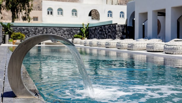 Una piscina con un moderno elemento acuático, un muro de piedra y un edificio con ventanas arqueadas al fondo.