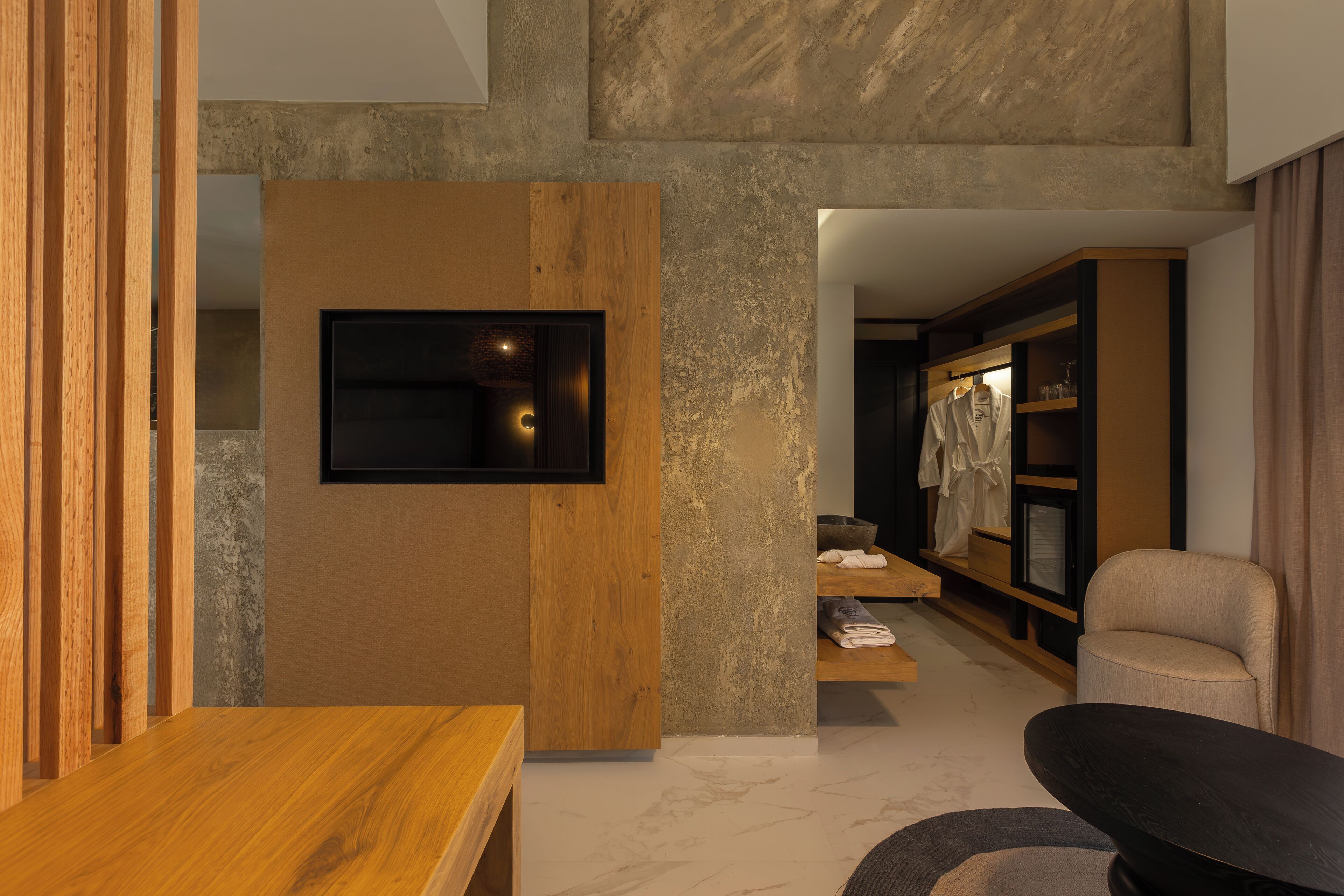 A modern living room with a wooden table, a flat-screen TV mounted on a textured wall, a comfortable armchair, and a built-in shelving unit with clothing and decorative items.