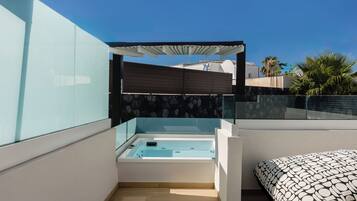 A modern balcony with a glass enclosure, a built-in hot tub, and a patterned sofa.