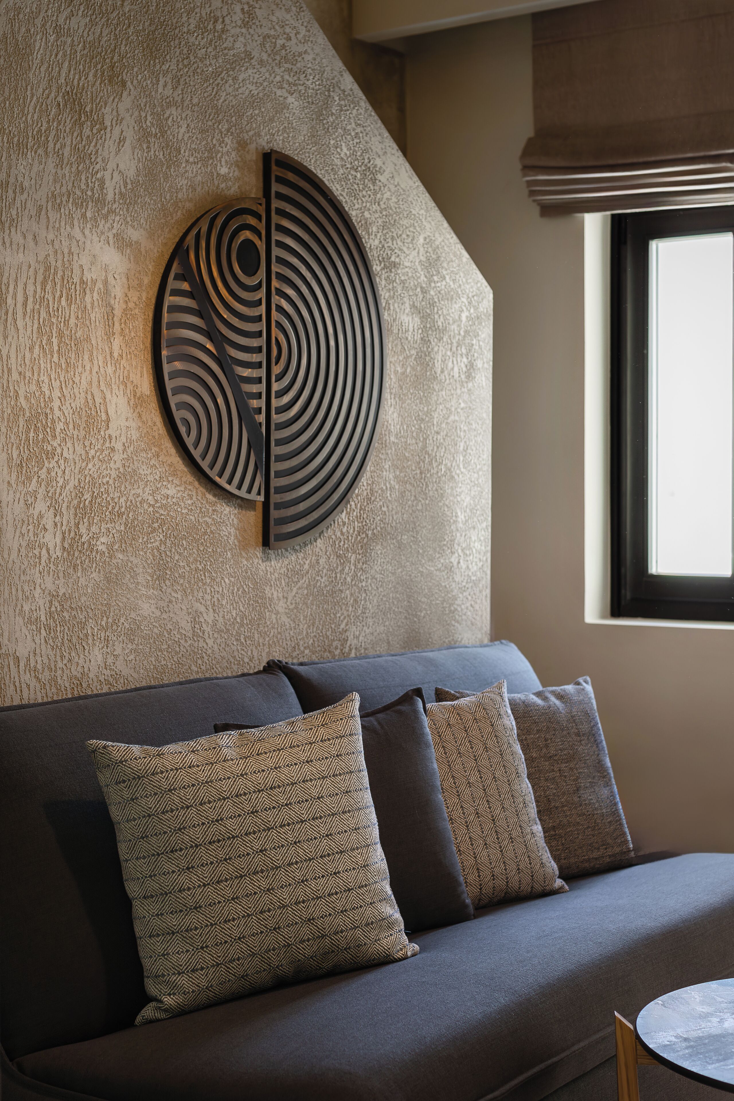 Una sala de estar moderna con una pared texturizada, una pieza de arte circular en la pared, un sofá oscuro con cojines estampados y una pequeña mesa redonda.