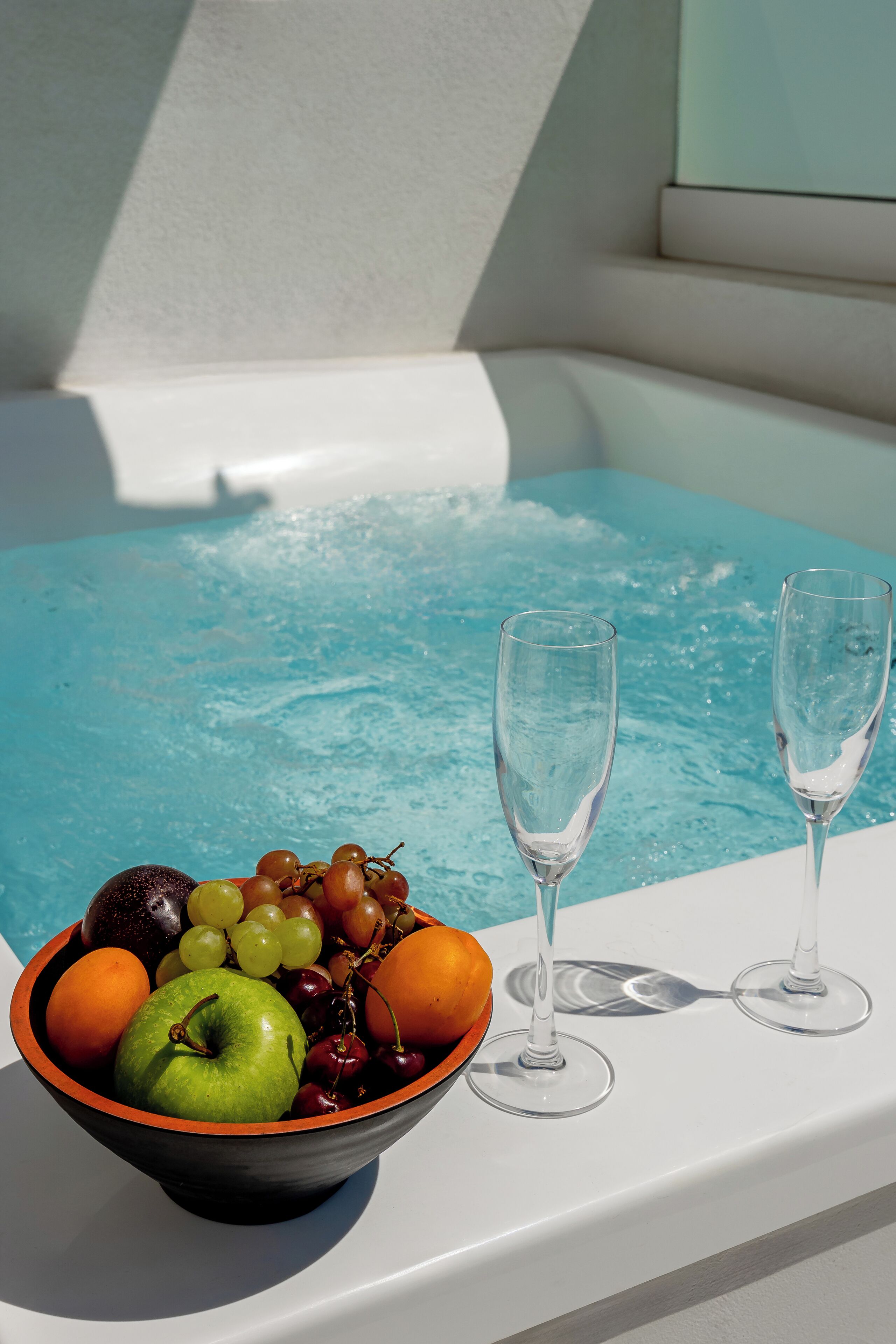A bowl of fruit and two glasses on a white surface by a pool.
