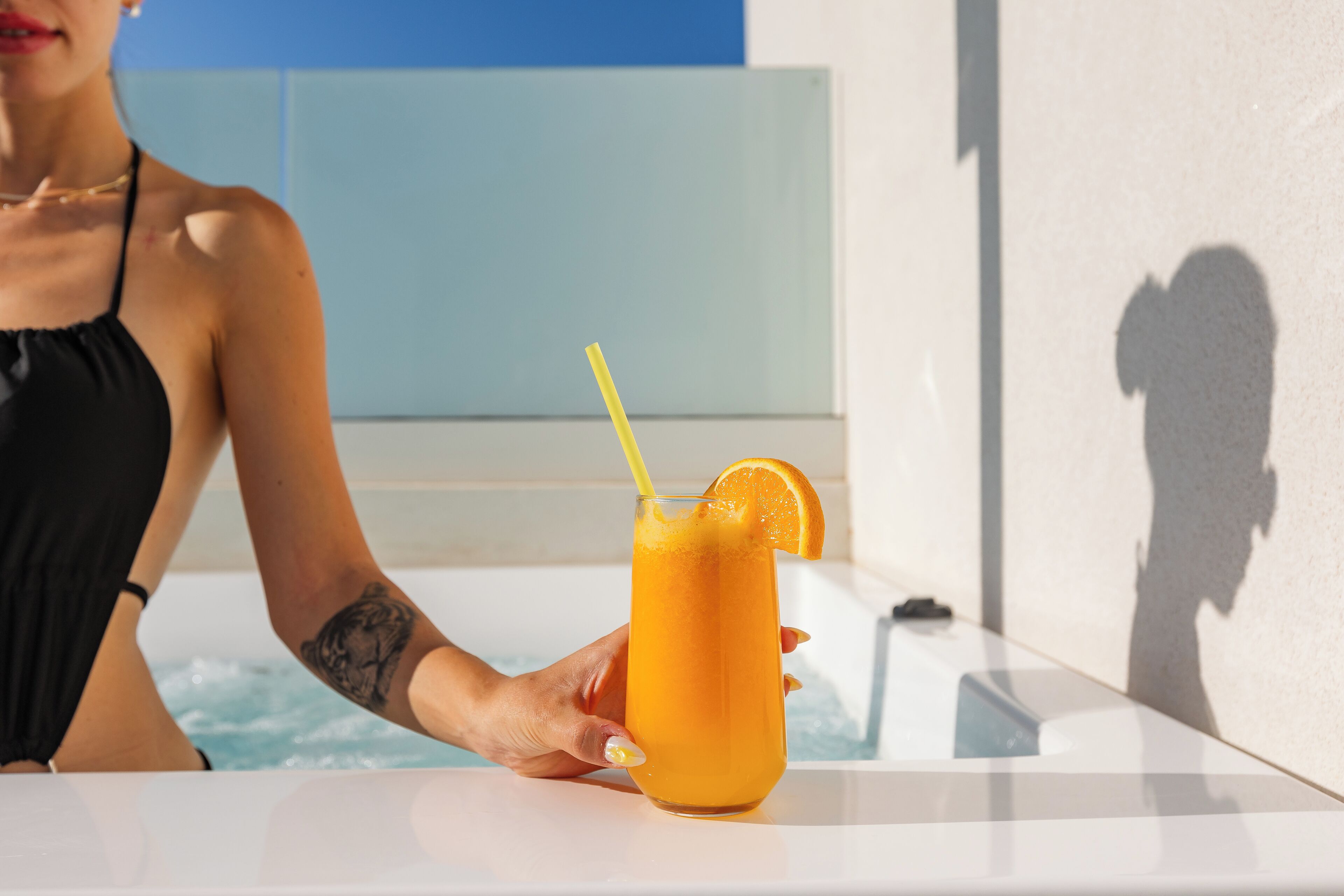 A person holding a tall glass with a yellow straw and an orange slice, near a pool area.