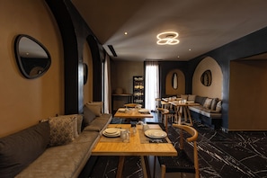 A modern dining area with a long wooden table set for a meal, a sofa, and a bar area with a wine rack in the background.
