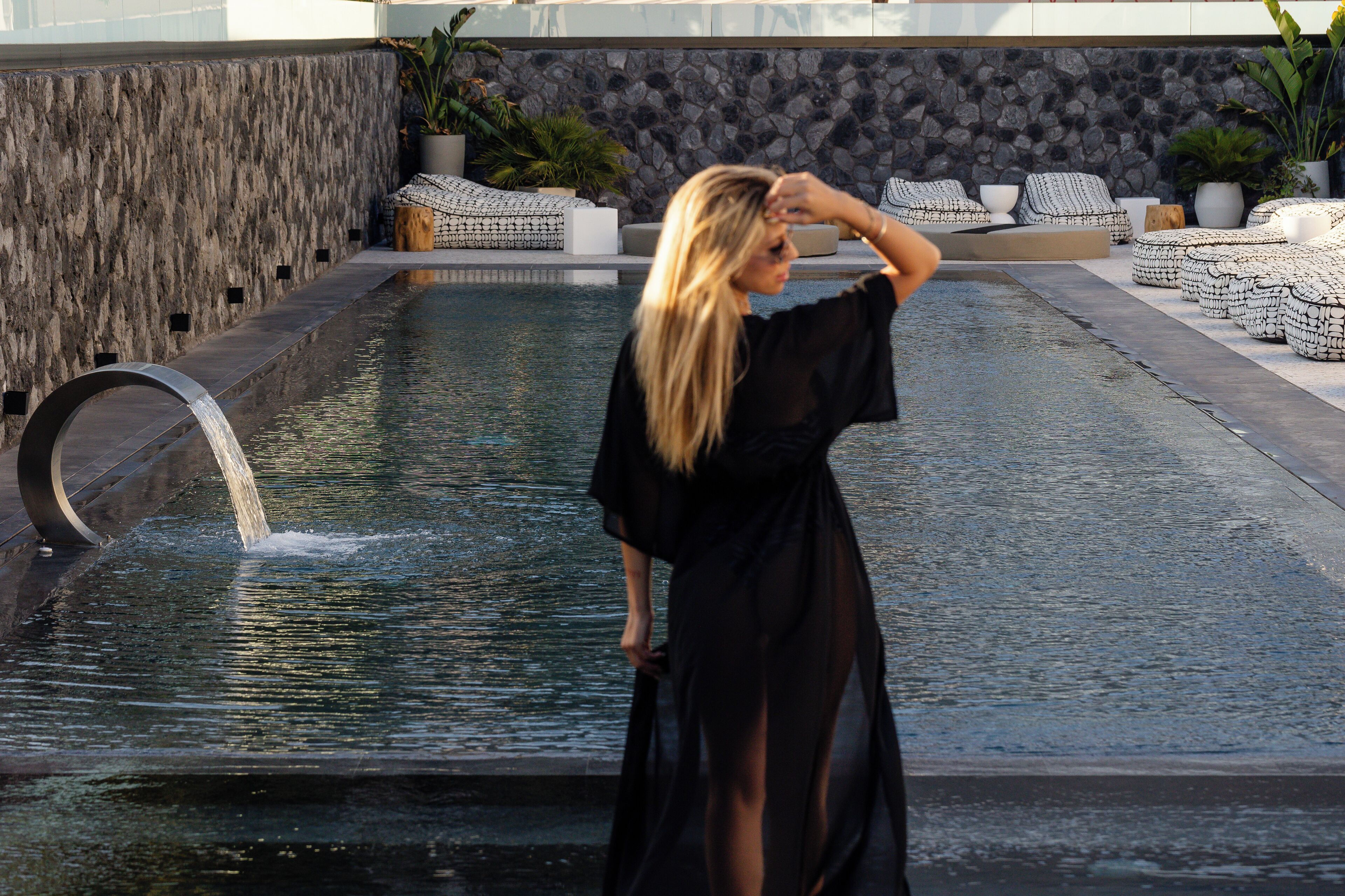 A woman in a black dress standing by a pool with a stone wall and seating area.