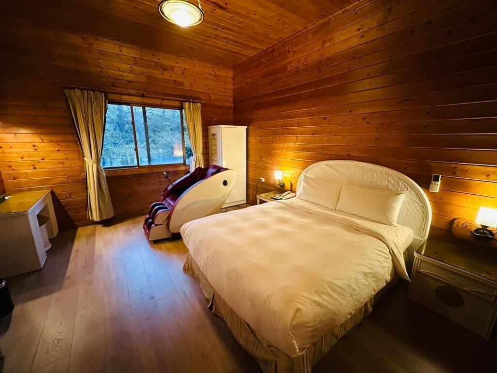 A wooden cabin bedroom with a bed, a desk, a chair, a refrigerator, and a window with curtains.