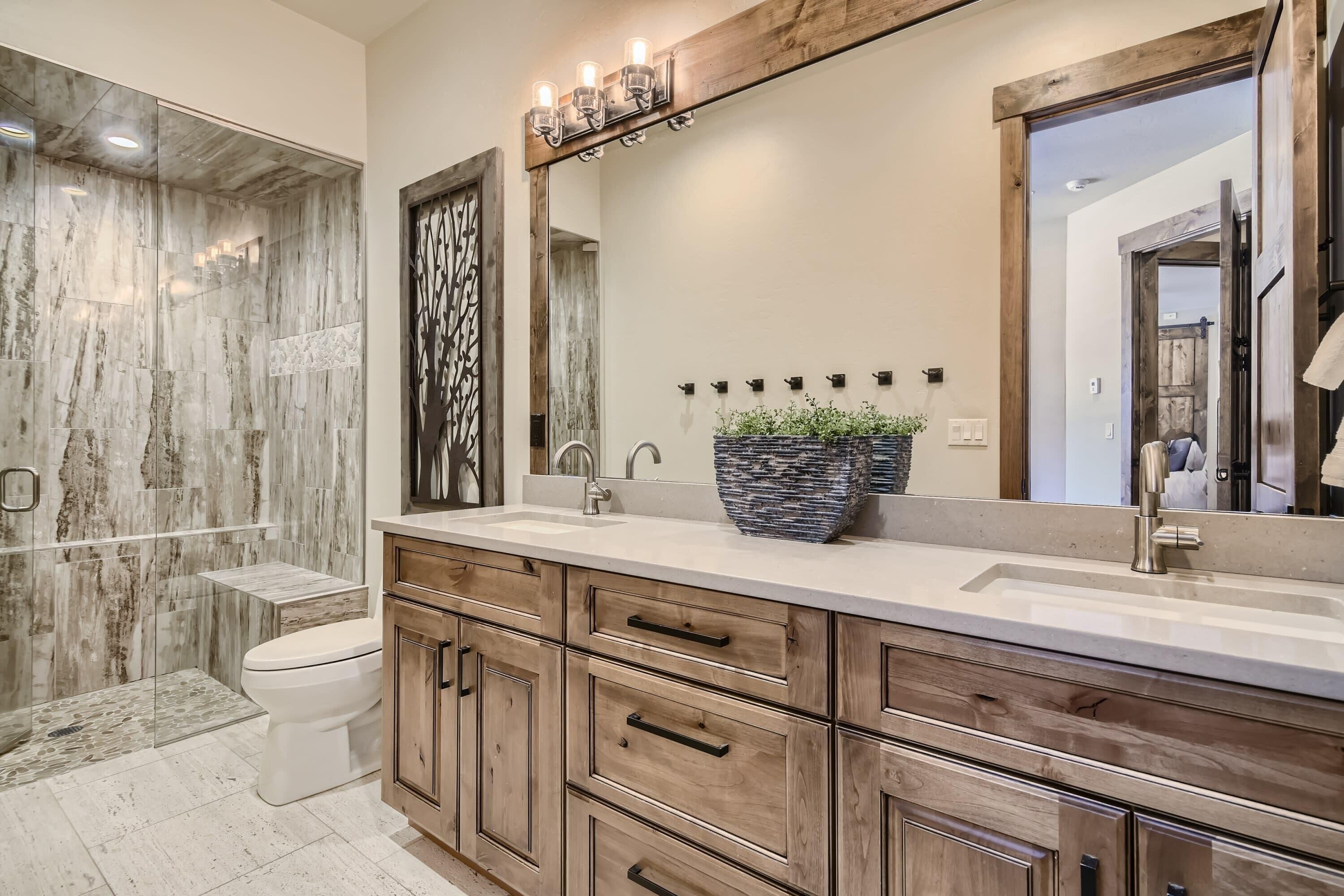 A bathroom with a marble shower wall, a wooden vanity with multiple drawers, a large mirror, and a potted plant.