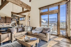 A spacious living room with a large window, a wooden coffee table, a leather sofa, and a kitchen area with wooden beams.
