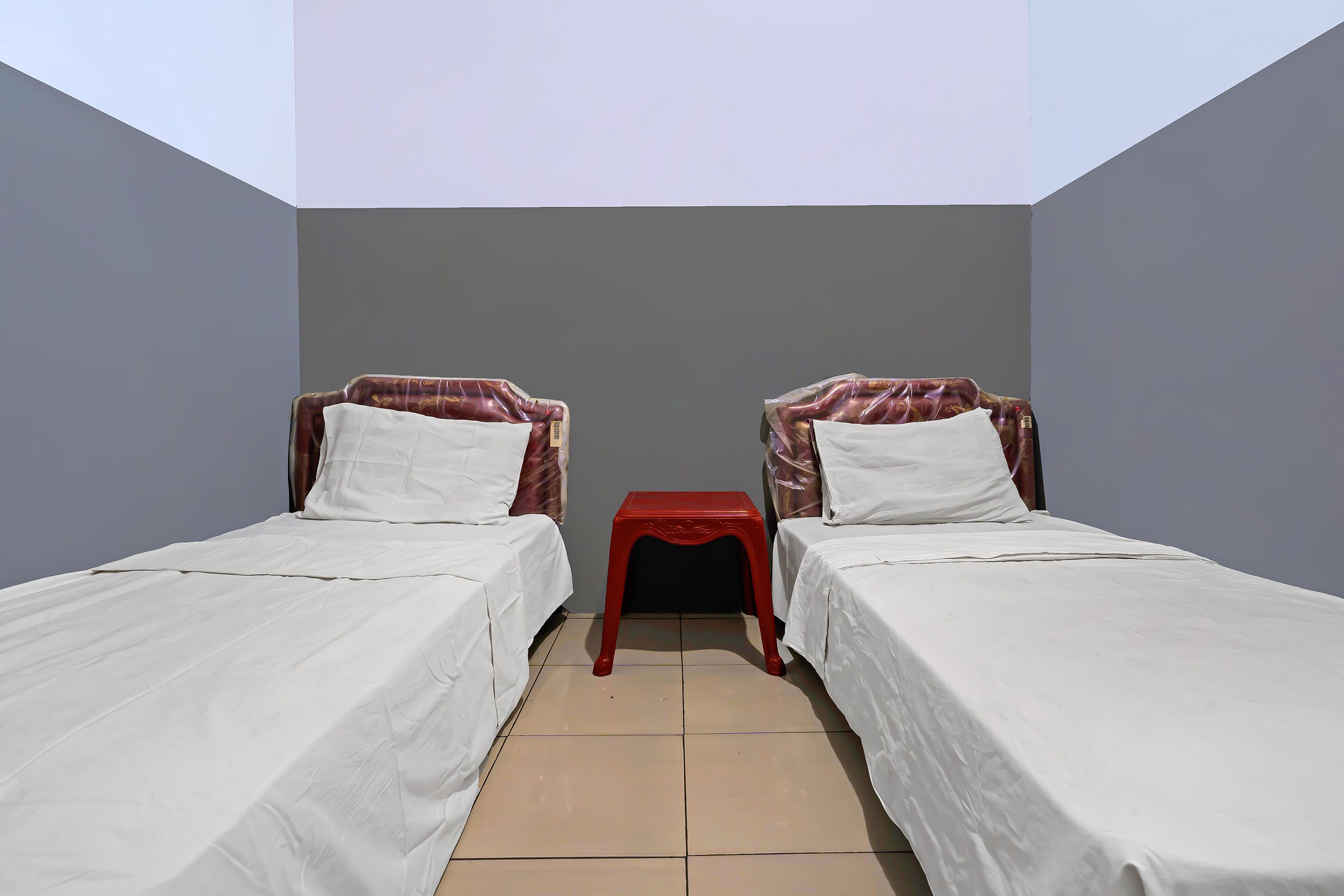 Ein kleines, sauberes Zimmer mit zwei Einzelbetten, einem kleinen roten Tisch und einer schlichten Wand.