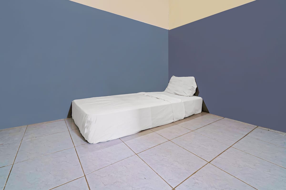 A small, single-bed room with a plain white bedspread and a pillow. The room has a simple layout with a tiled floor and plain walls in blue and beige tones.