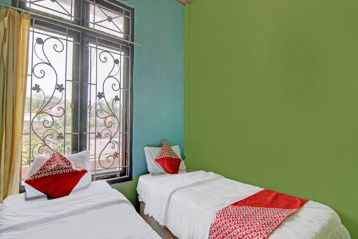 Une chambre avec deux lits, chacun agrémenté de draps blancs et d’oreillers rouges, une fenêtre ornée de barreaux en fer décoratifs et un mur vert.