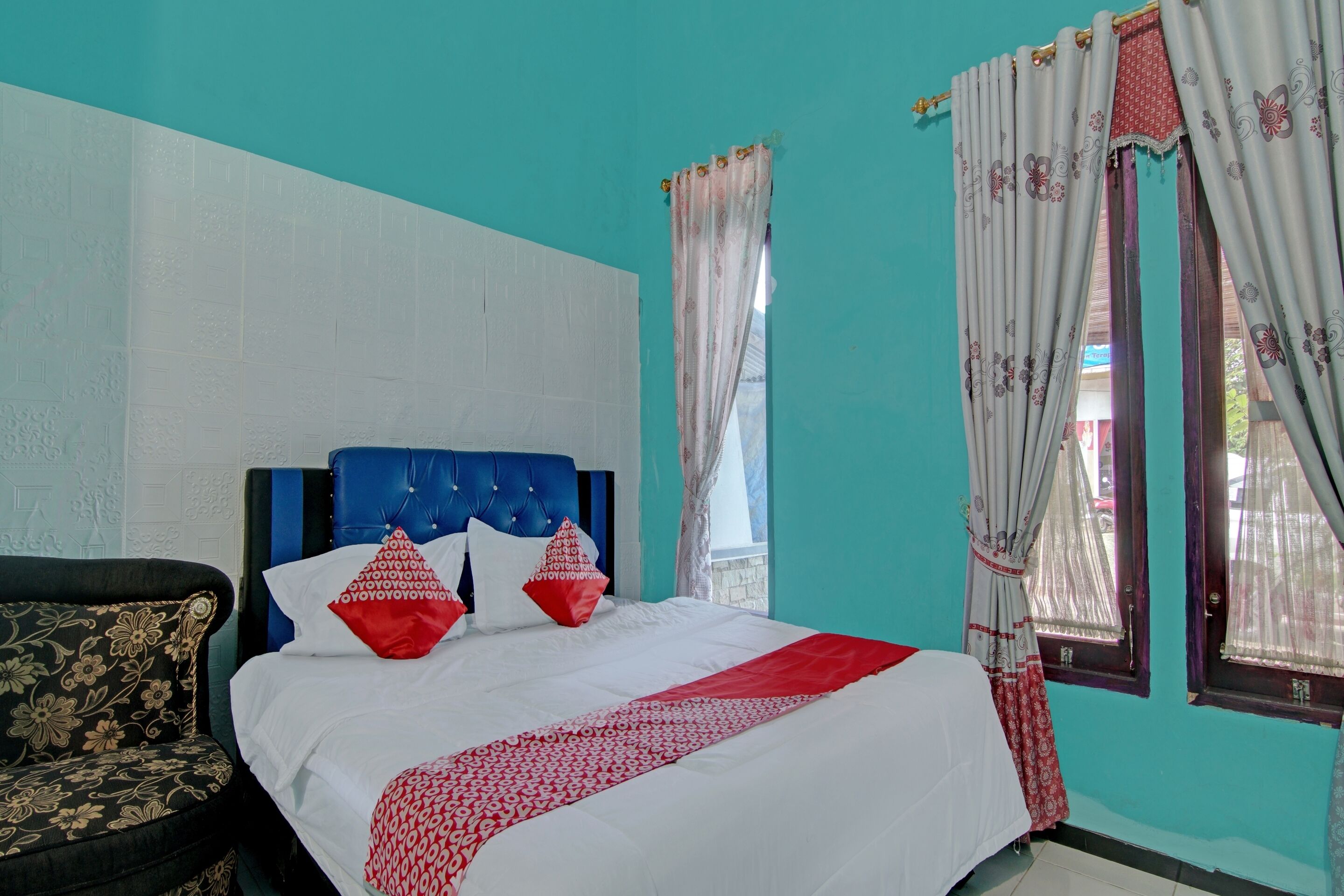 Quarto com cabeceira azul, cama com roupa de cama branca e vermelha, sofá floral e janela com cortinas.