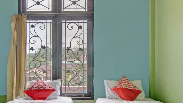 Twee enkele bedden met witte lakens en kussens met een rood-wit patroon, een raam met decoratieve metalen tralies en uitzicht op groen buiten.