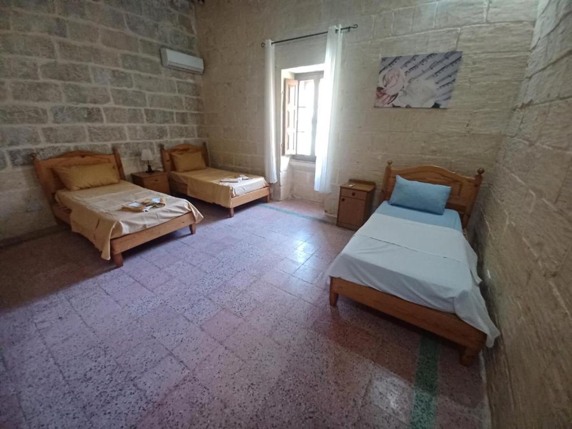 A room with three wooden beds, a stone wall, a window with curtains, and a small table.
