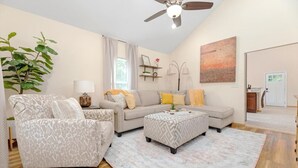 A living room with a sofa, armchair, ottoman, and a ceiling fan. There is a plant, a lamp, and a painting on the wall.