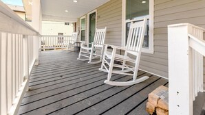 A covered porch with white rocking chairs and a table, wooden deck, and a house with beige siding.