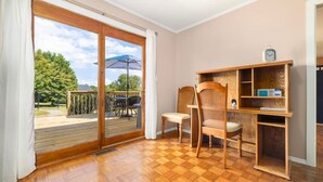 Een kamer met een houten vloer, een schuifdeur naar een terras, twee rieten stoelen, een houten boekenkast en een klok aan de muur.