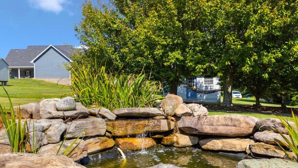 Een vijver met rotsen en een kleine waterval, omringd door groen en een huis op de achtergrond.